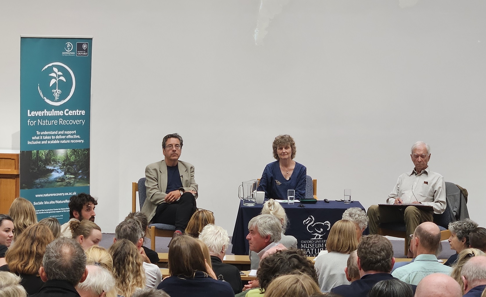 A banner for the Leverhulme Centre for Nature Recovery sits on the stage inhabited by George Monbiot, EJ. Milner-Gulland, and Allan Savory.