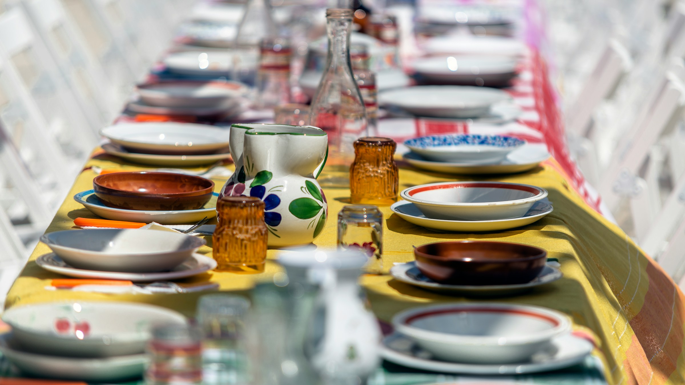 A long table full of colorful empty plates and cups on a colorful tablecloth flanked by white folding chairs. Photo by Mario Caruso via Unsplash.