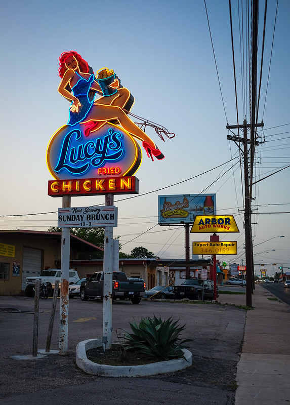 A photo of a roadside neon sign for "Lucy's Fried Chicken" featuring a woman in a dress and high heels clutching a tub of fried chicken. Photo by Lars Plougmann via Flickr.