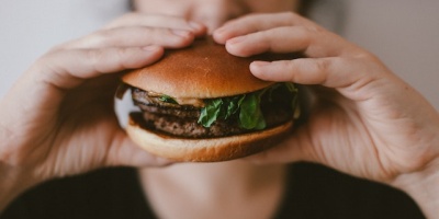 A person lifts a burger towards their face which is out of focus behind the burger. Photo by Szabó Viktor via Unsplash.