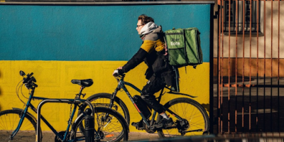 Uber Eats delivery person on an ebike rides past a wall painted like the Ukrainian flag. Photo by Ammy Singh via Pexels.