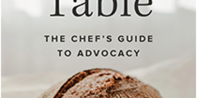 The cover of At the Table featuring a loaf of bread on a white tablecloth