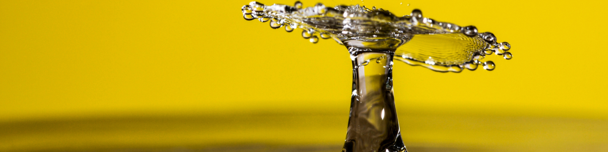A droplet of water hits the surface in freeze frame on a yellow background.