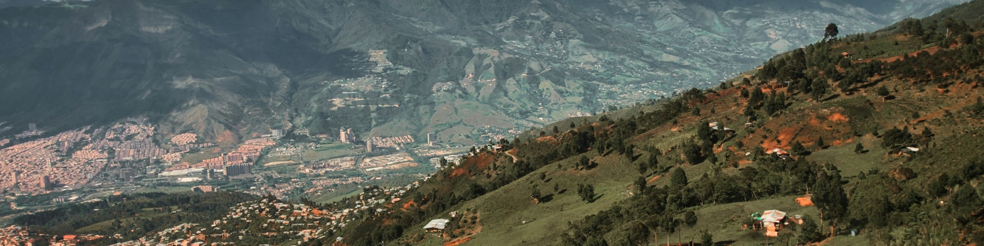 A hillside in Medellín with houses sprawled in the valley in the distance and mountains beyond that. Photo by Jan Brennenstuhl via UnSplash.