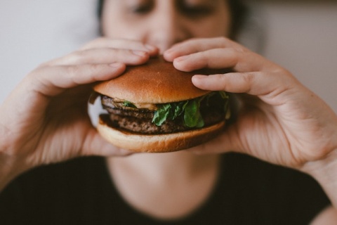 A person lifts a burger towards their face which is out of focus behind the burger. Photo by Szabó Viktor via Unsplash.