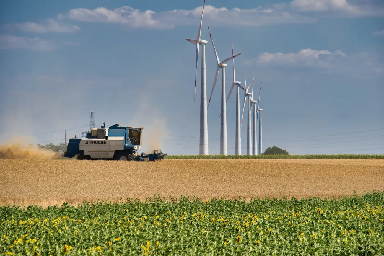 A combine harvests wheat in the foreground with a row of turbines in the distance. Photo by Anton Klyuchnikov via Pexels.