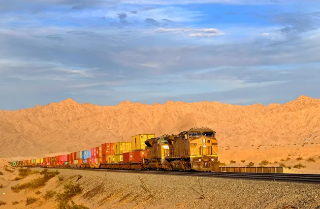 A train carrying many shipping containers through a desert landscape. Photo by jdblack via Pixabay.