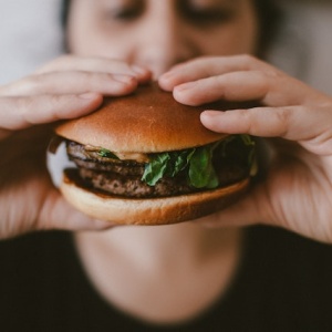 A person lifts a burger towards their face which is out of focus behind the burger. Photo by Szabó Viktor via Unsplash.