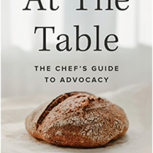 The cover of At the Table featuring a loaf of bread on a white tablecloth
